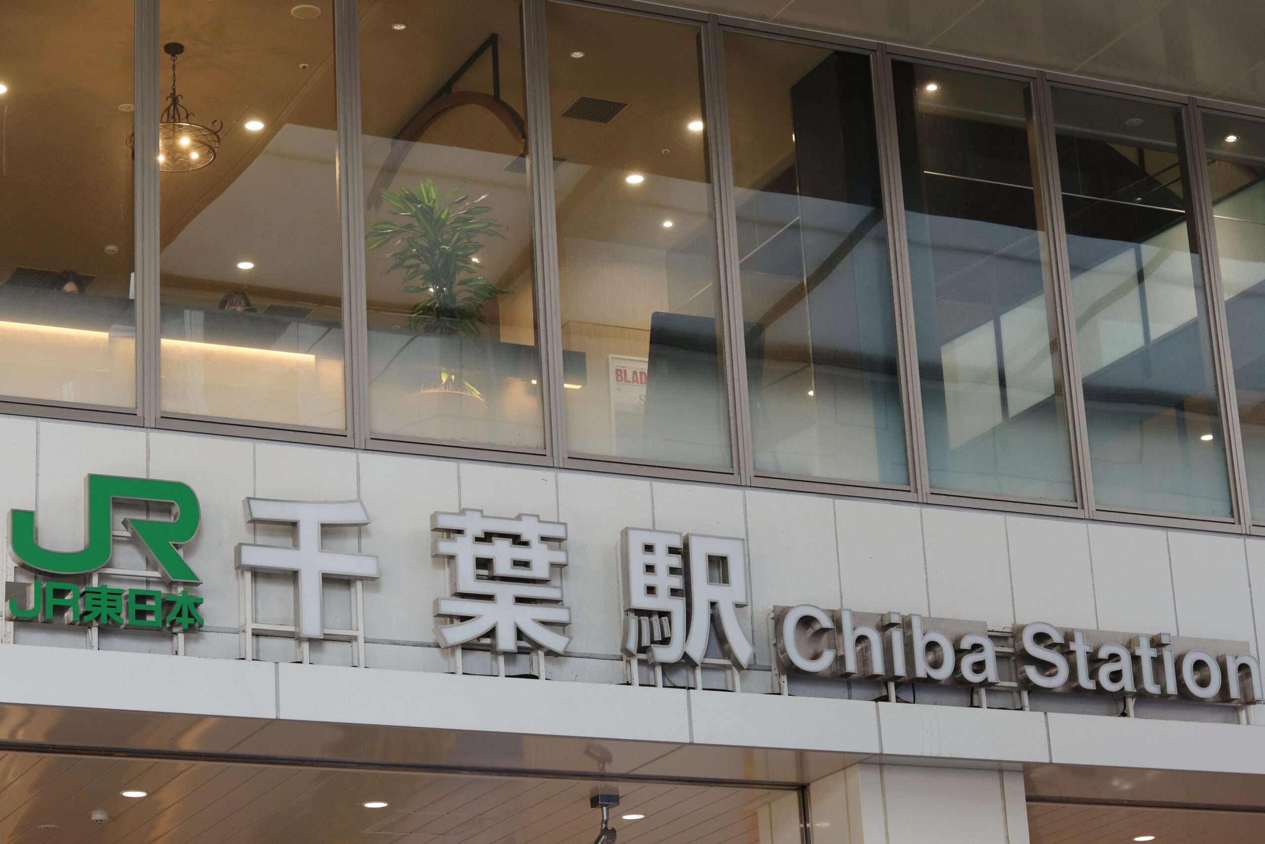 千葉駅の入り口には日本語と英語の標識が飾られ、ガラス窓からは植物のある内部の部屋を覗くことができます。近くには韓国語教室の存在を示唆する標識があり、駅の文化的多様性にさらに彩りを添えています。