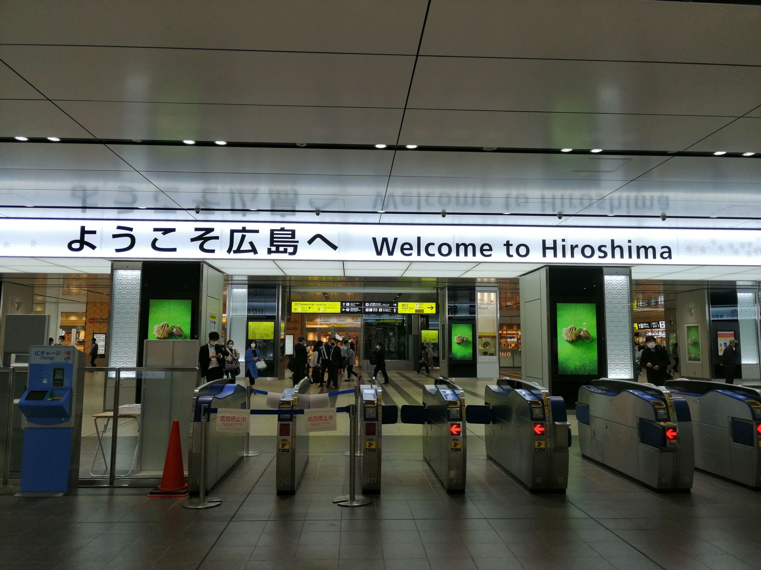 英語、日本語、韓国語教室で「ようこそ広島へ」と書かれた看板が掲げられた駅入口。広島の活気に満ちた中心地の広々とした天蓋の下にある改札口を、旅行者が慌ただしく通り抜けていきます。