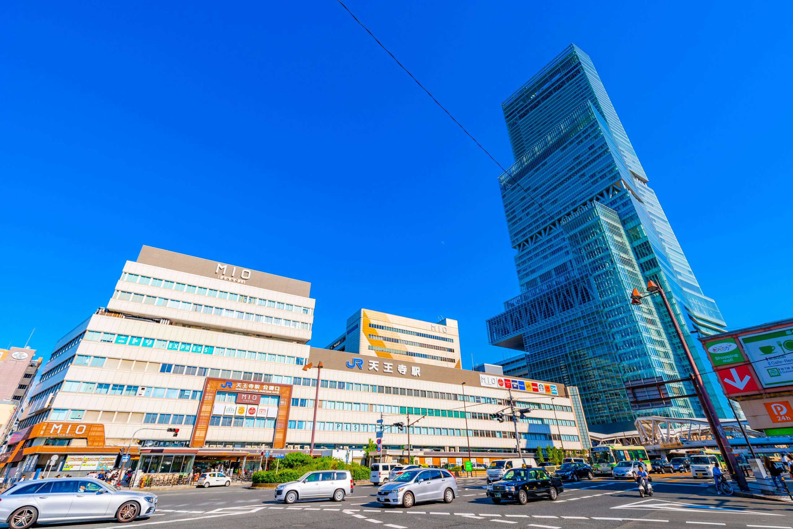 天王寺の都会的な風景は、澄んだ青空の下、大きな近代的なビルや高層タワーの前に車が行き交う賑やかな交差点を特徴としています。近くには、環境に豊かな文化を加えた活気に満ちた韓国語教室があります。