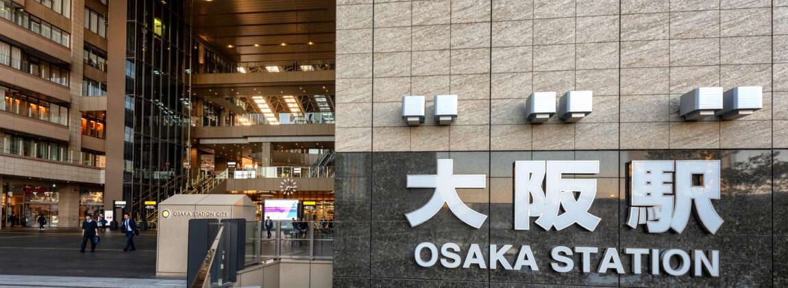 この画像は大阪駅の入口を捉えたもので、壁には駅名が大きく表示されている。近くでは、韓国語のレッスンを提供する語学学校「大阪韓国語教室」の広告の前を通り過ぎる人々がいる。