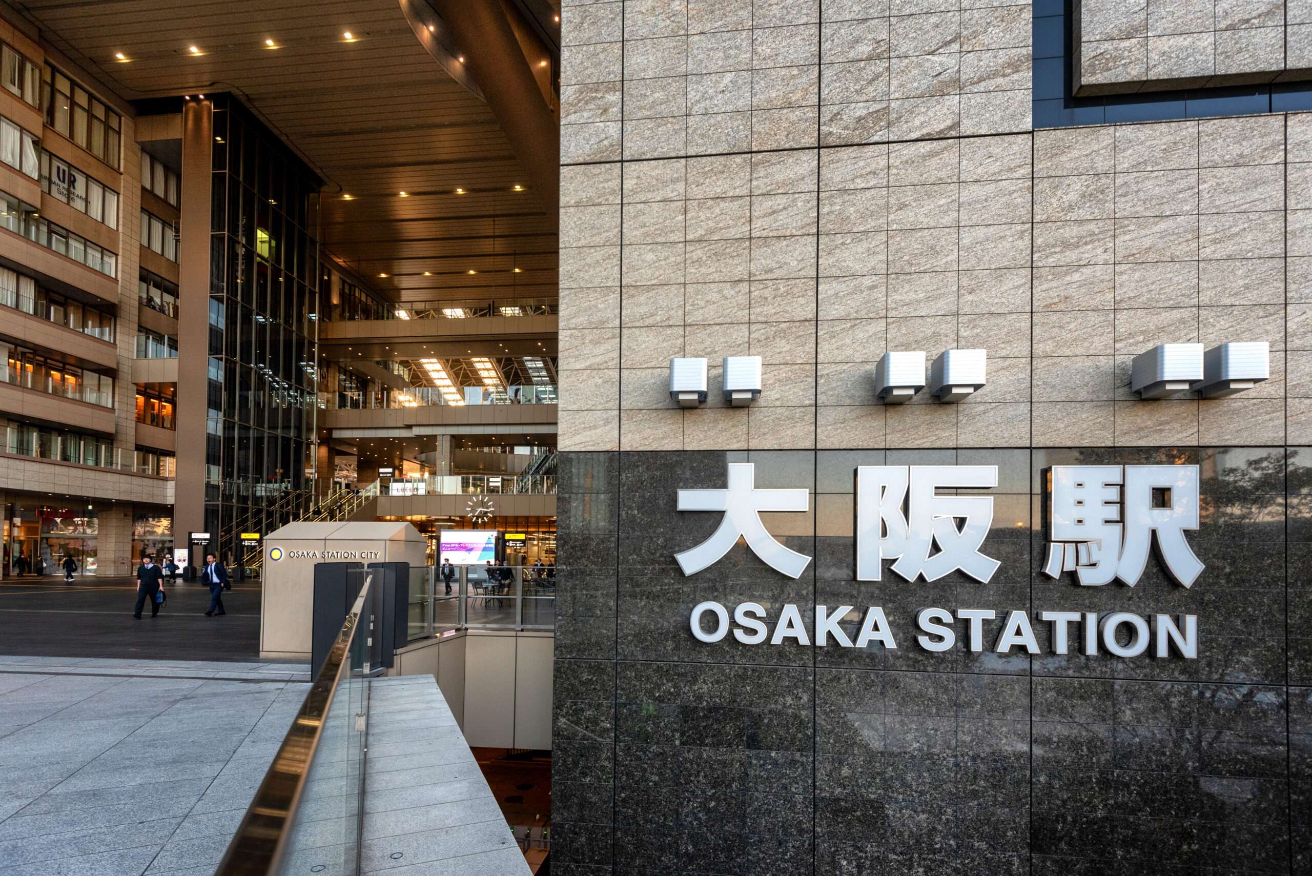 この画像は大阪駅の入口を捉えたもので、壁には駅名が大きく表示されている。近くでは、韓国語のレッスンを提供する語学学校「大阪韓国語教室」の広告の前を通り過ぎる人々がいる。