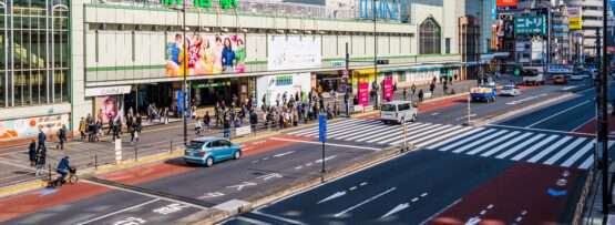 新宿の賑やかな市街地のストリートビュー。車、自転車、歩行者が「JR」と「ルミネ」と書かれた大きな建物の近くを行き交い、韓国語を話す人々の活発な会話が都市生活の活気あるタペストリーに彩りを添えています。