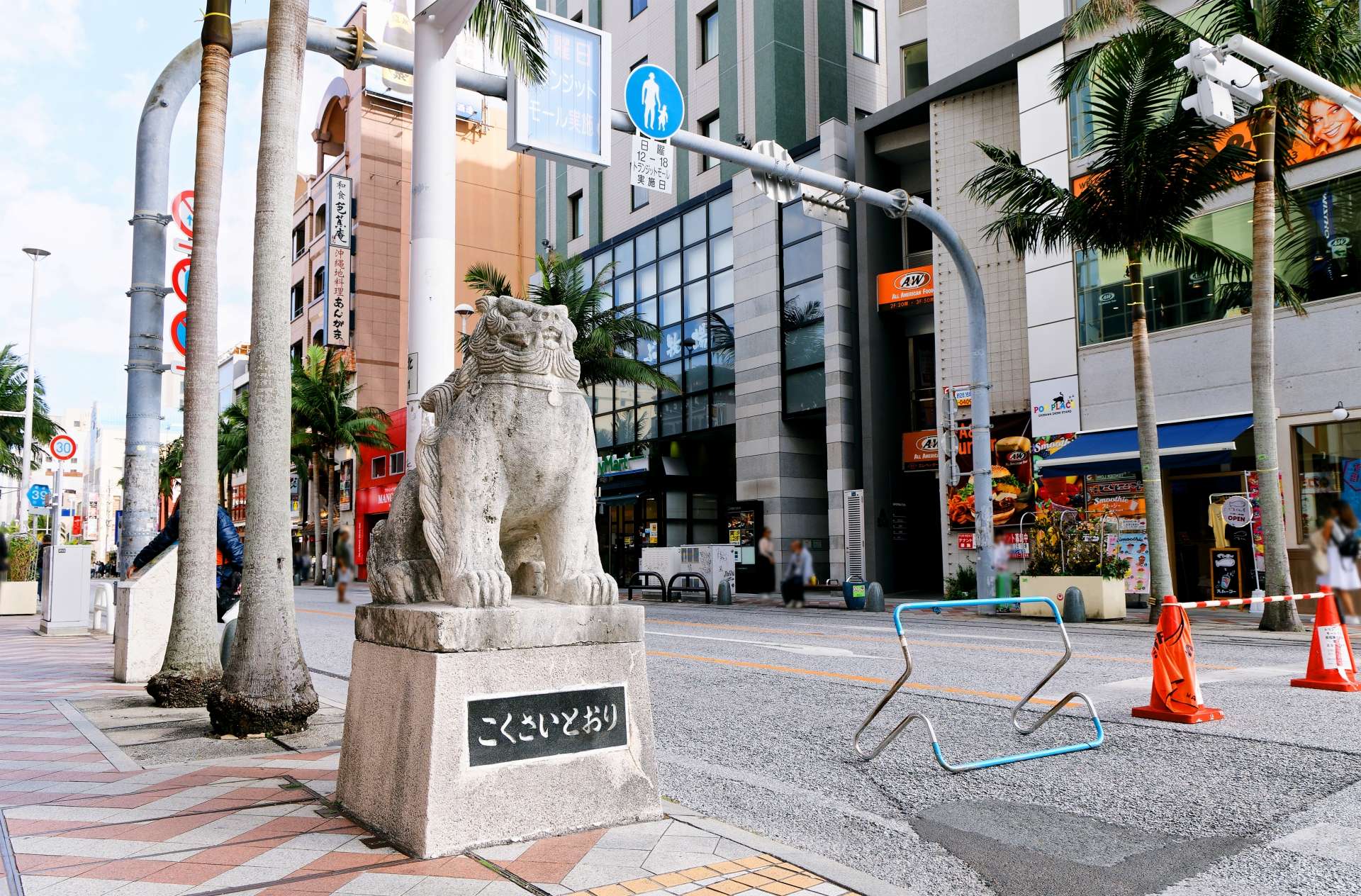 ヤシの木が優しく揺れ、交通標識が通行人を誘導する中、沖縄・那覇のにぎやかな街角にライオンの石像が立っている。近くの韓国語教室では、活気に満ちた都会の中で語学レッスンを行っています。
