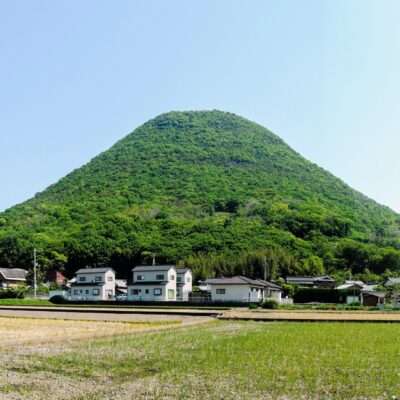 香川・高松の小さな村の背後には、青々とした円錐形の丘がそびえ立ち、前景には澄んだ青空の下、田畑が広がっています。近くの韓国語教室では、好奇心旺盛な住民に文化交流や語学レッスンを提供しています。