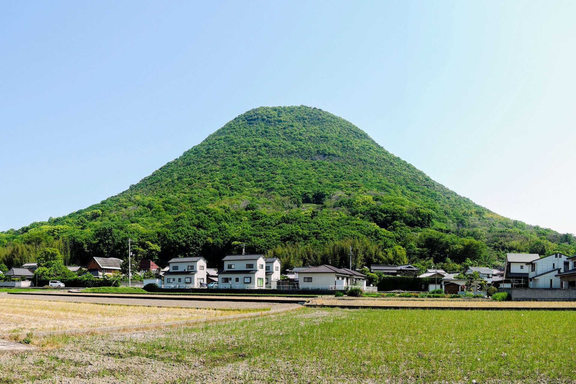 香川・高松の小さな村の背後には、青々とした円錐形の丘がそびえ立ち、前景には澄んだ青空の下、田畑が広がっています。近くの韓国語教室では、好奇心旺盛な住民に文化交流や語学レッスンを提供しています。
