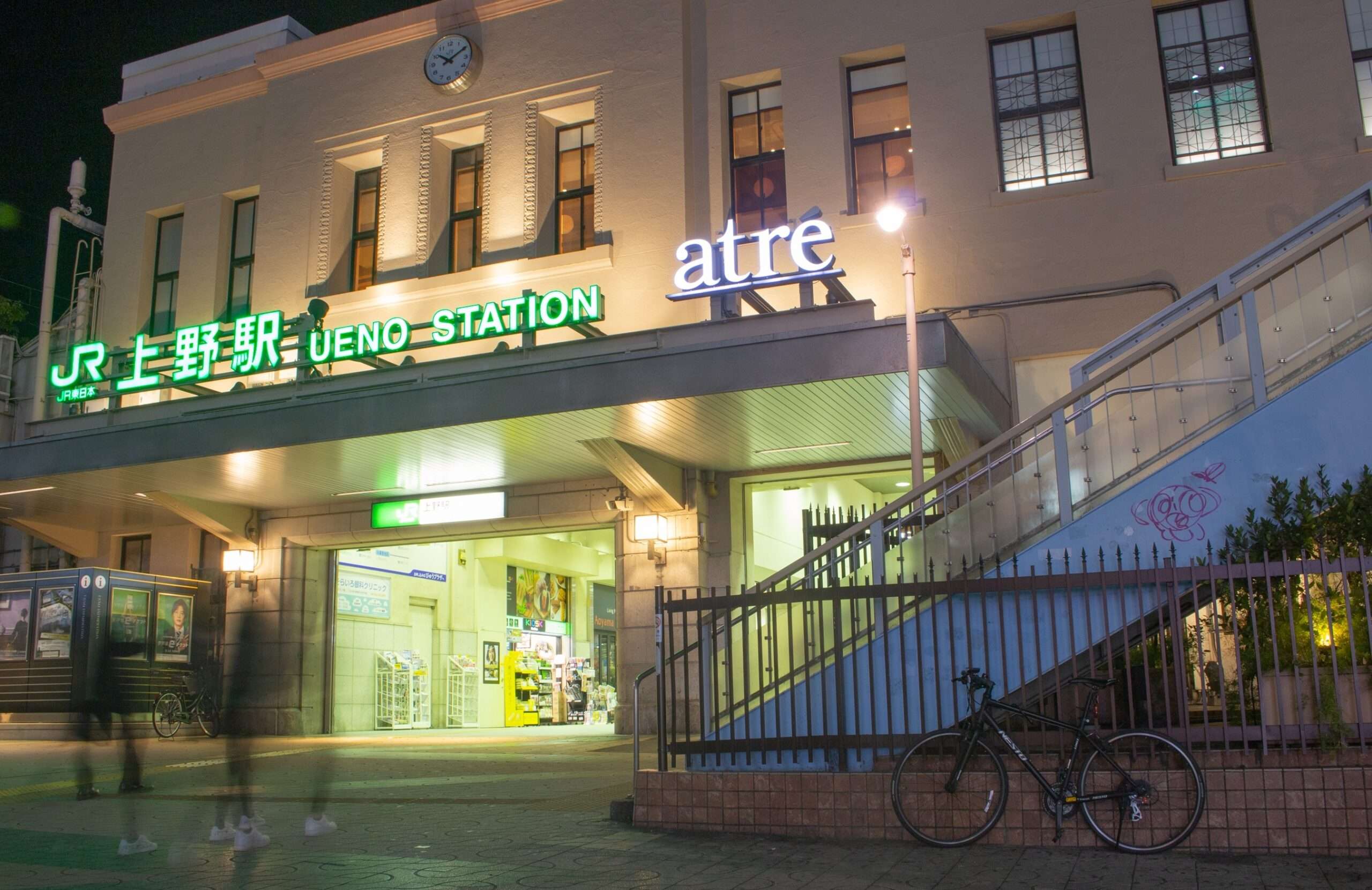 夜の上野駅の外には光る看板があり、近くには自転車が停められている。人々がぼんやりと動いている。この人気の通勤ハブの外では、喧騒が続いている。近くには、韓国語を学ぶために言語愛好家が集まる韓国語教室がある。