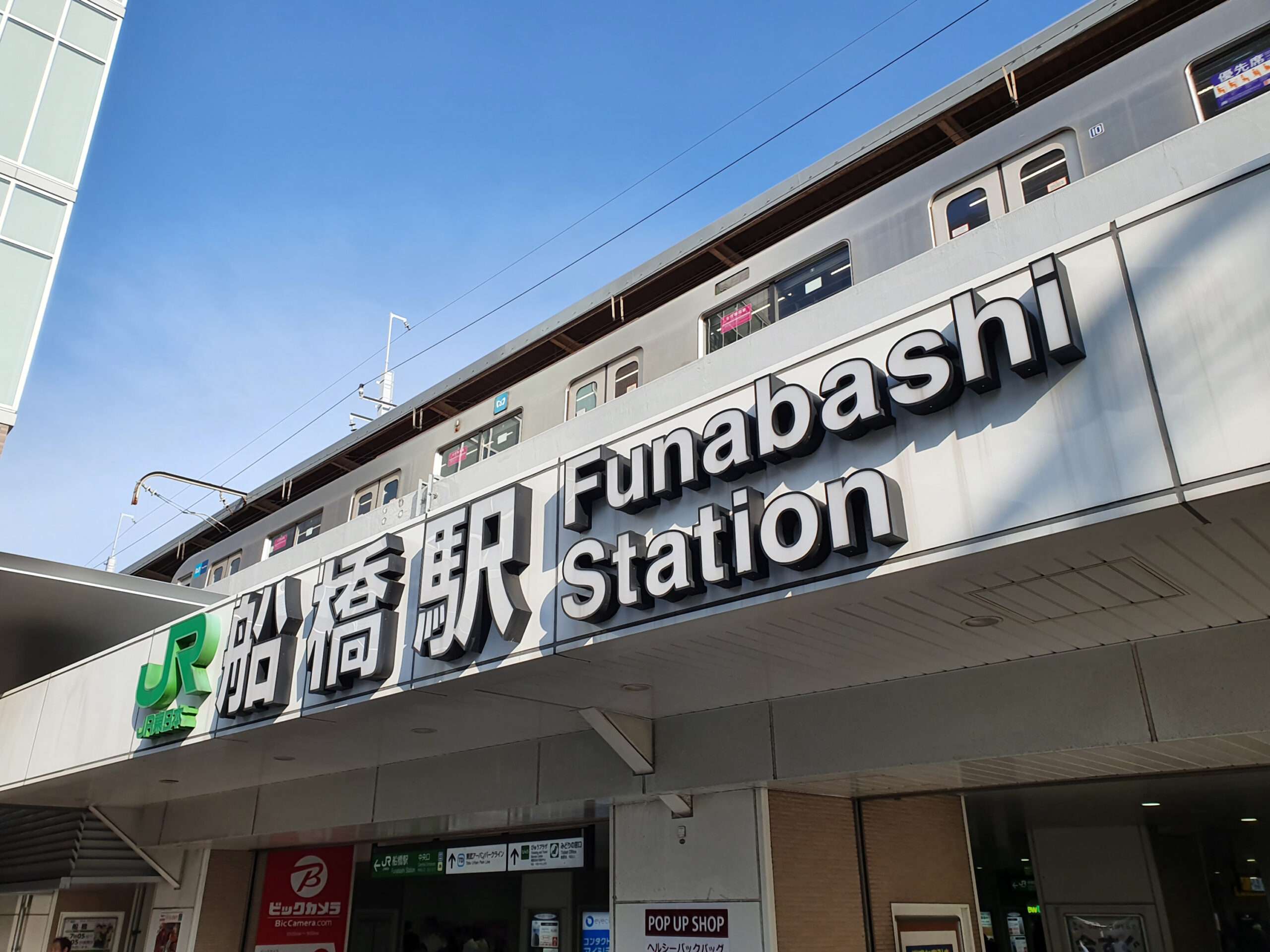 船橋駅の正面には、活気ある文化の融合を暗示する日本語と英語の大きな看板が掲げられています。入り口の上には電車が部分的に見えます。近くには、韓国語と韓国文化の体験を提供する韓国語教室があります。