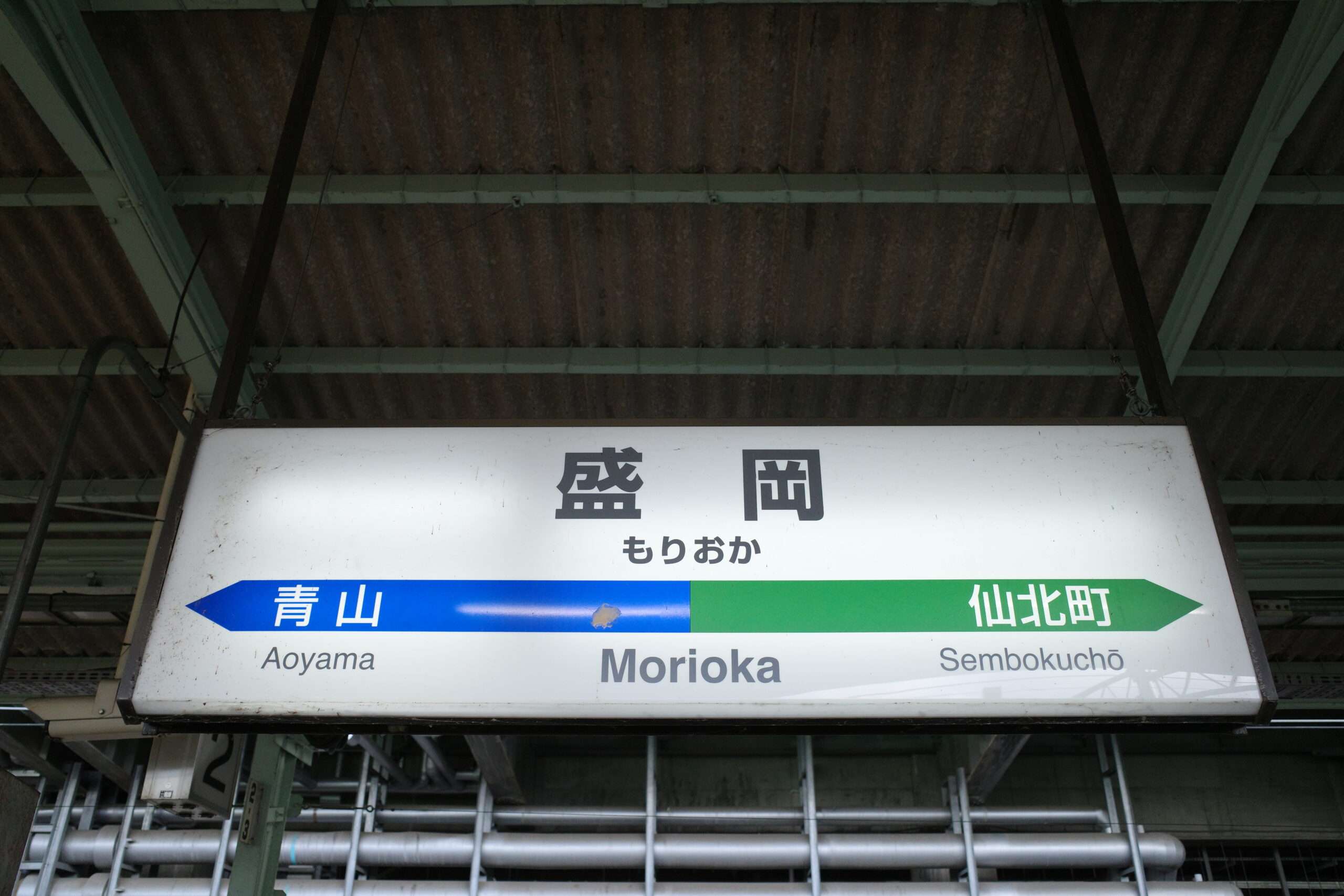 盛岡駅では、金属パイプと波形の屋根を背景に、旅行者に青山と仙北町への案内標識が設置されています。岩手・盛岡にあるこの環境は、地元の魅力と近くの韓国語教室のような国際的な機会を融合させています。