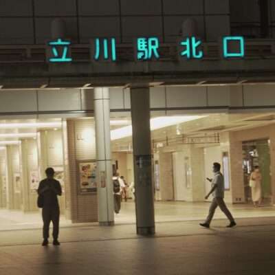 夜、立川駅の入り口付近を歩く人々。中には携帯電話に夢中になっている人もいる。駅のネオンサインが上に輝き、近くにある韓国語教室の広告が目を引く。