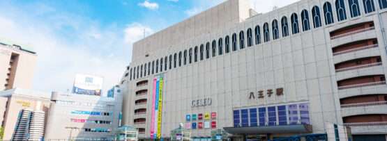 日本の八王子駅の外観。看板のあるベージュ色の大きな建物、陸橋、澄んだ青い空が特徴。近くには、この活気に満ちた街並みの中で韓国語を学びたい人のための韓国語教室があります。