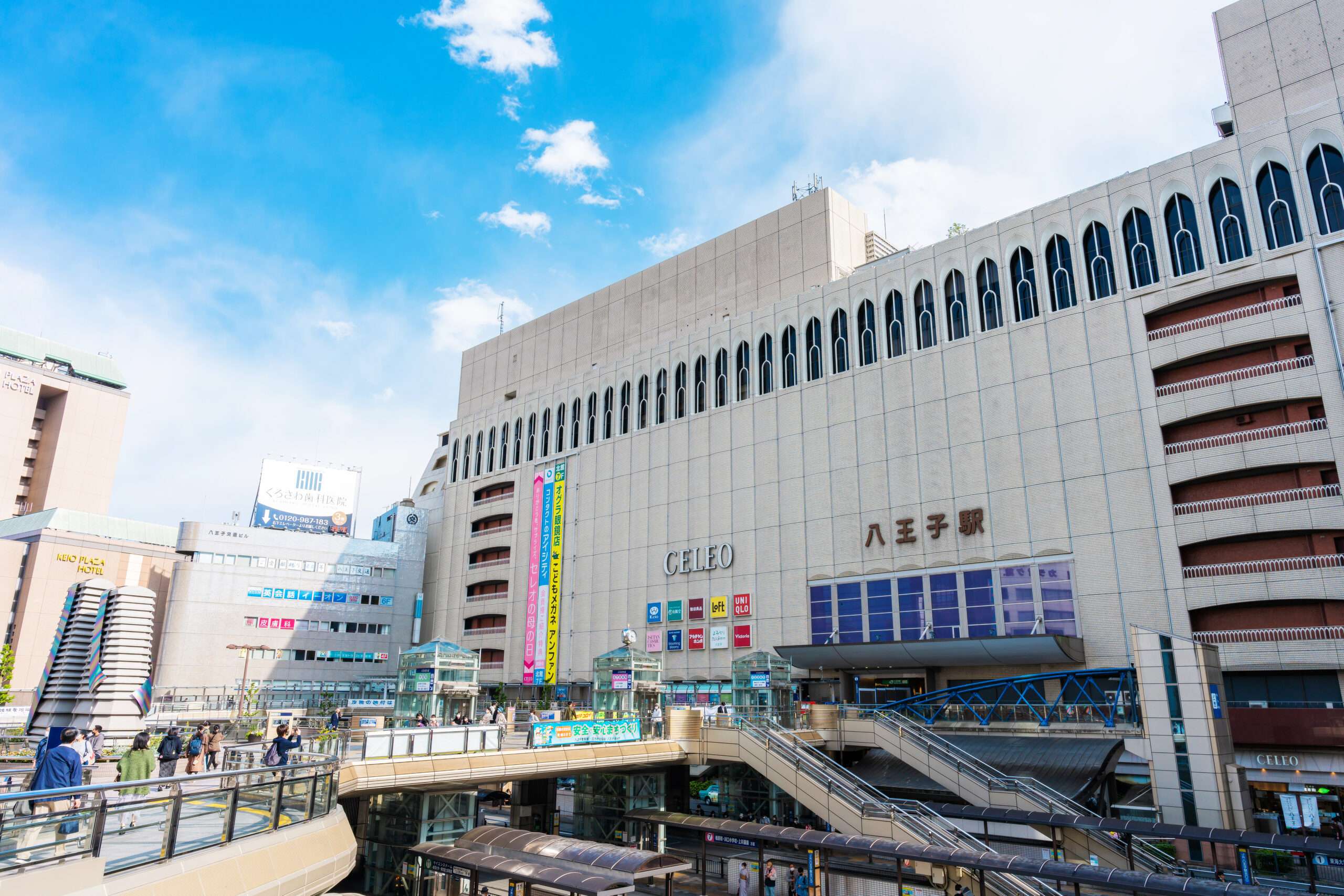 日本の八王子駅の外観。看板のあるベージュ色の大きな建物、陸橋、澄んだ青い空が特徴。近くには、この活気に満ちた街並みの中で韓国語を学びたい人のための韓国語教室があります。