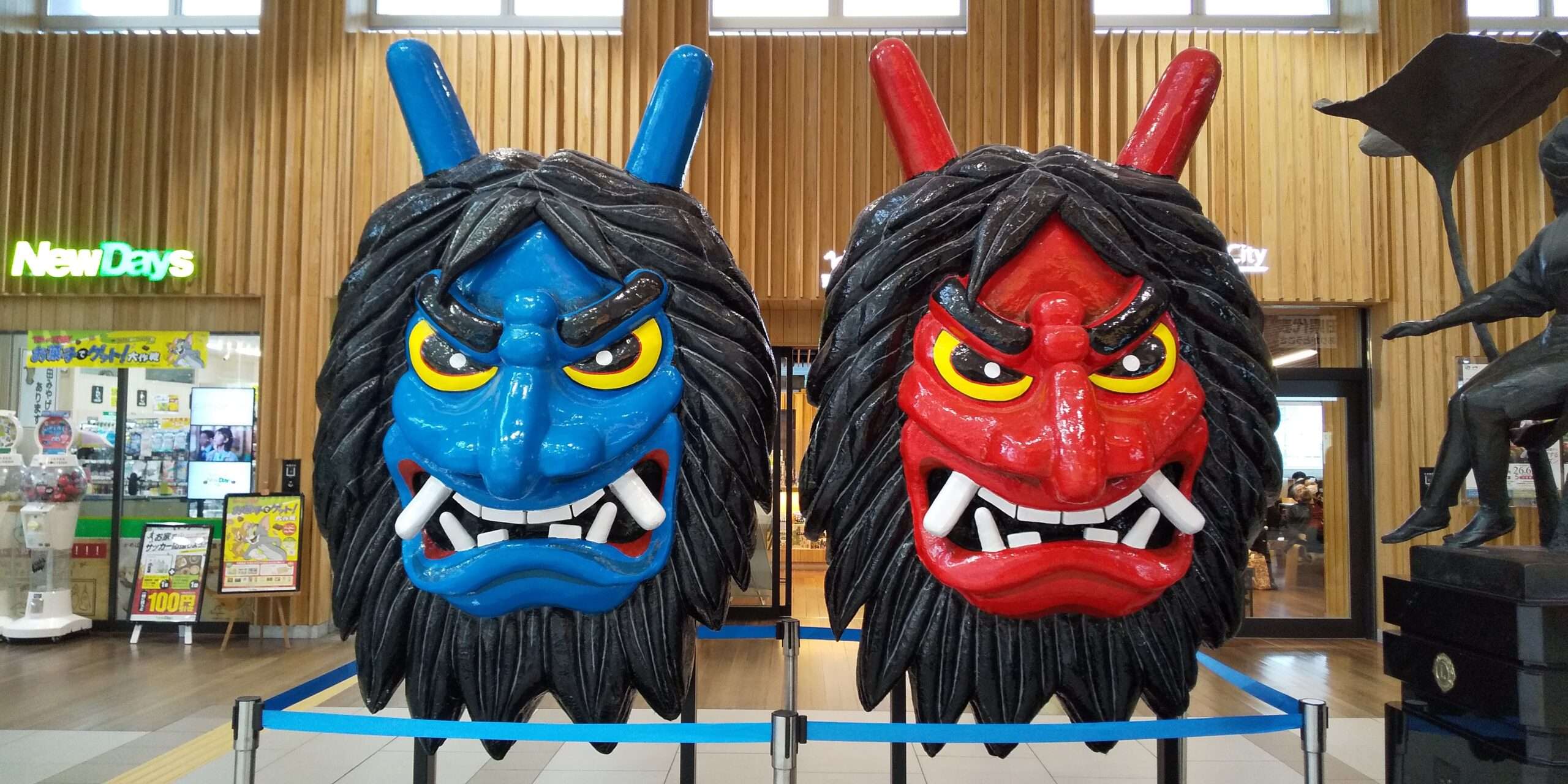 角と牙が付いた大きくて色鮮やかな鬼の面が二つ（一つは青、一つは赤）ガラス窓近くの木の壁を背景に屋内に展示されており、秋田の展示会で見られるような鮮やかな文化遺物を彷彿とさせます。