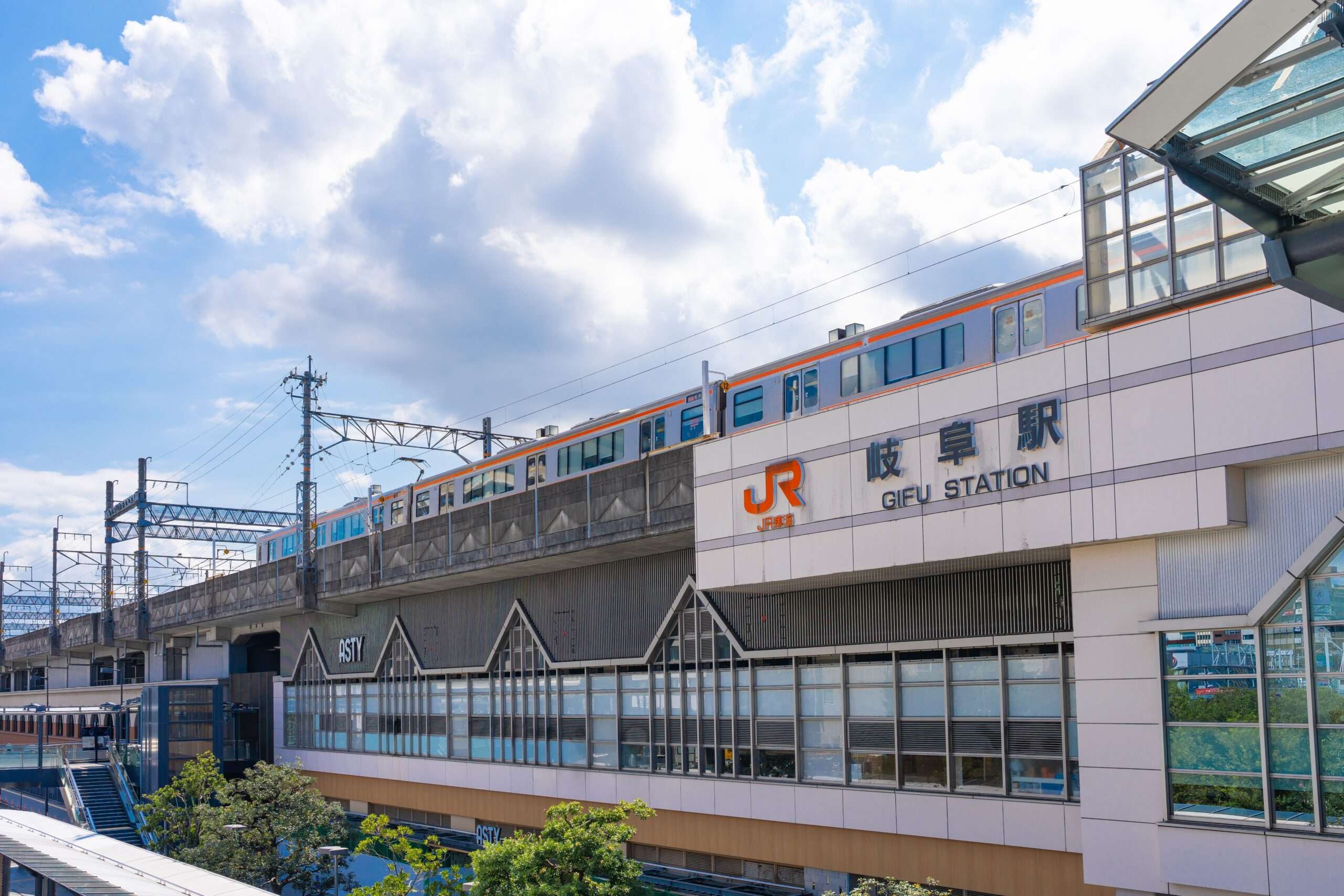 薄曇りの空の下、日本語と英語の標識が掲げられた駅舎の前を電車が通過していく。近くにある韓国語教室では、岐阜の多様な文化を紹介しています。