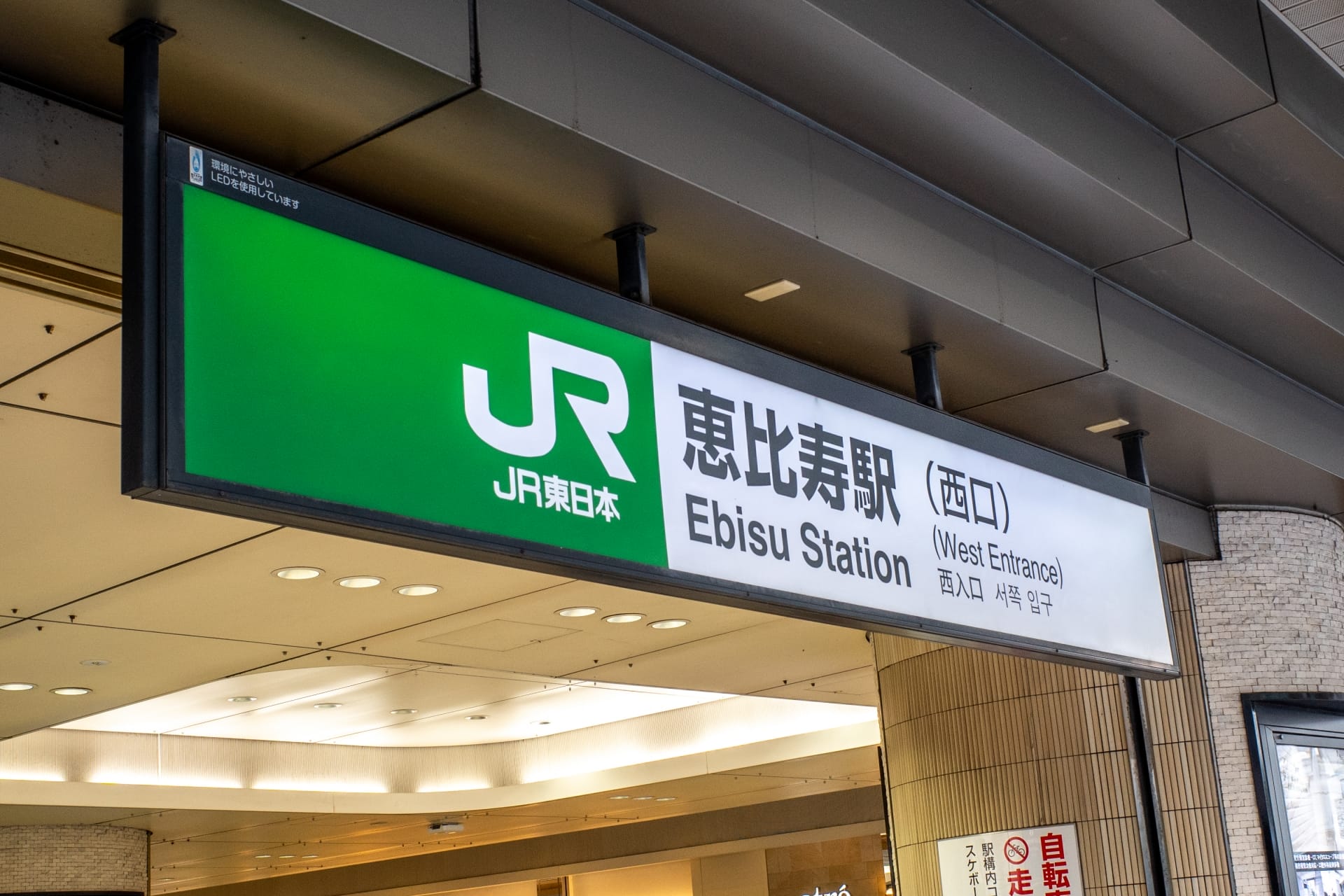 東京の恵比寿駅西口の看板。建物の天蓋の下に JR 東日本のロゴと場所の詳細が記載されています。近くには、個人向けの韓国語レッスンに興味のある方向けの韓国語教室マンツーマンもあります。.