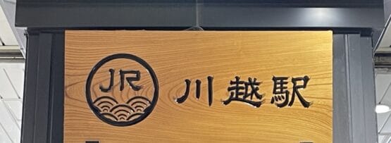 日本語の文字とJRのロゴが入った木製の看板が川越駅の入り口にあり、旅行者を賑やかな中心地へと誘います。.