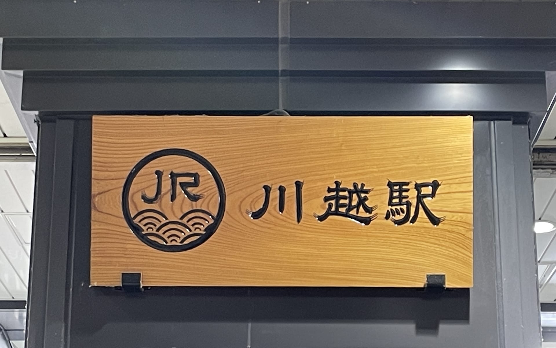 日本語の文字とJRのロゴが入った木製の看板が川越駅の入り口にあり、旅行者を賑やかな中心地へと誘います。.