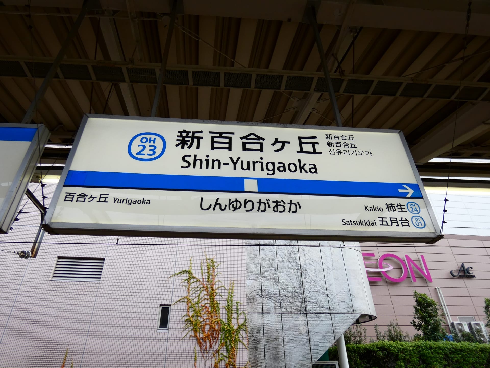 新百合ヶ丘 (新百合ヶ丘) の駅標識。行き方、プラットフォーム番号 OH 23、および左側に百合ヶ丘、右側に近くの駅が記載されています。背景に韓国語教室マンツーマンの建物の一部が見えます。.