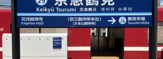 京急鶴見駅の看板。青い空の下、背景にホームと赤い電車が見えます。韓国語学習におすすめの教室からほど近い場所にあります。.