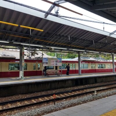 金沢八景駅のホームでは、反対側で栗色とクリーム色の電車が待っています。数人がベンチの横に集まり、おそらく次回の韓国語教室マンツーマンのセッションについて話し合っているのだろう。線路の向こうを見つめる彼らを、頭上の屋根が守ってくれます。.