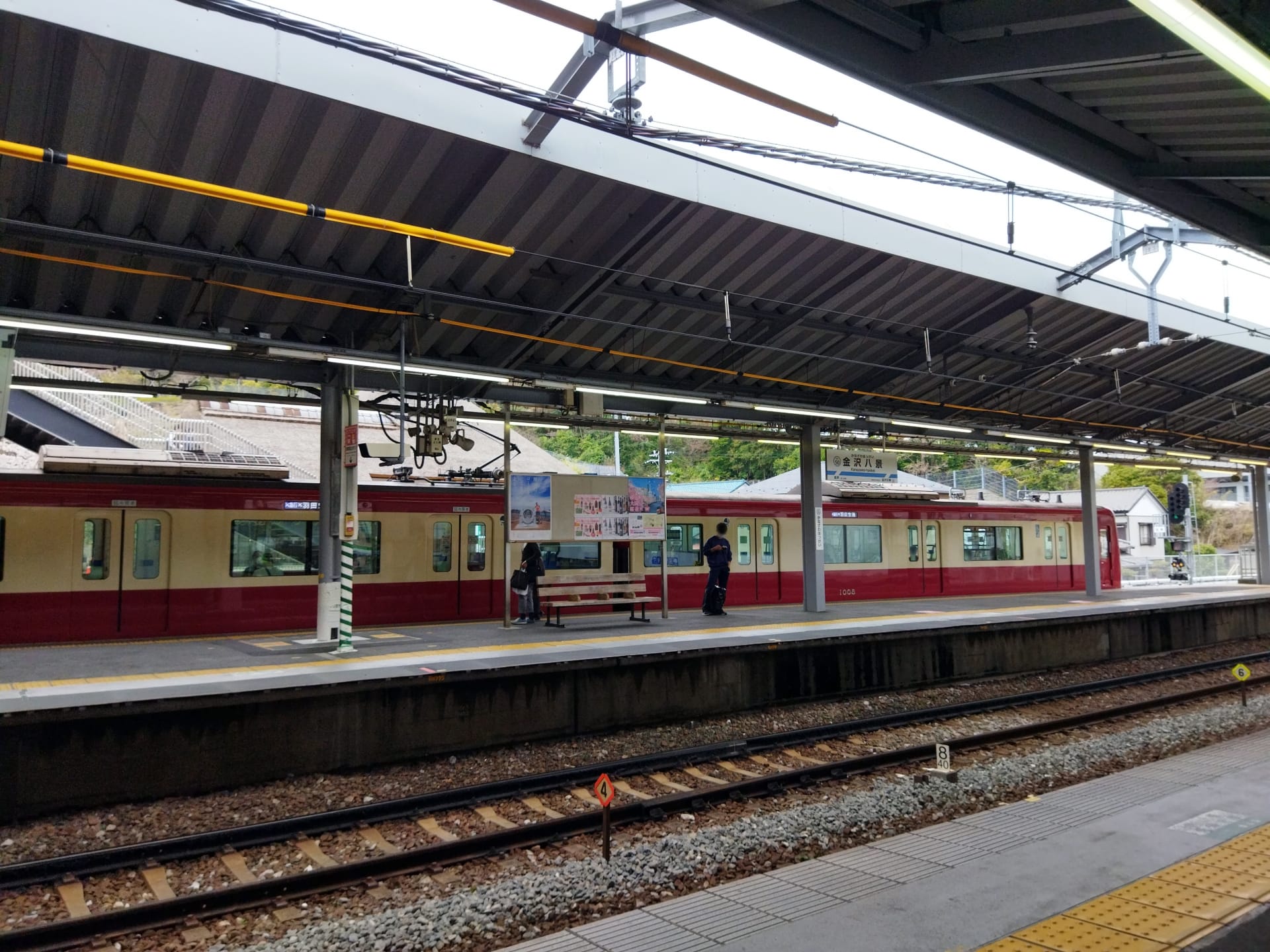 金沢八景駅のホームでは、反対側で栗色とクリーム色の電車が待っています。数人がベンチの横に集まり、おそらく次回の韓国語教室マンツーマンのセッションについて話し合っているのだろう。線路の向こうを見つめる彼らを、頭上の屋根が守ってくれます。.