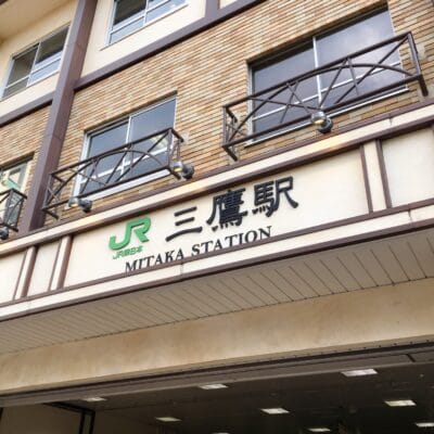 三鷹駅の入り口には日本語と英語の標識があり、木造の建物の外観にある象徴的な JR のロゴの下に、近くにある韓国語教室マンツーマンの詳細も表示されています。.