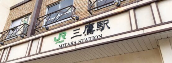 三鷹駅の入り口には日本語と英語の標識があり、木造の建物の外観にある象徴的な JR のロゴの下に、近くにある韓国語教室マンツーマンの詳細も表示されています。.