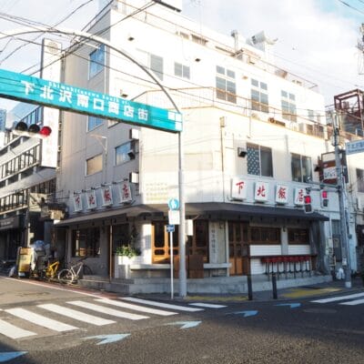 下北沢の街並みには、青い日本語の看板の下にある建物や店舗が特徴です。自転車は左側にあり、交差点は信号機で見渡せます。一方、韓国語教室マンツーマンの広告が、この魅力的な近所のスナップショットに文化的な雰囲気を加えています。.