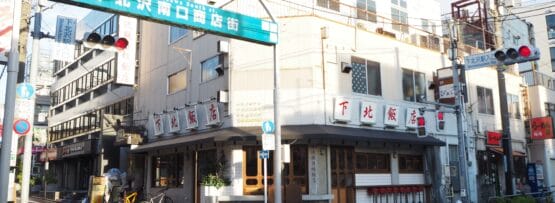 下北沢の街並みには、青い日本語の看板の下にある建物や店舗が特徴です。自転車は左側にあり、交差点は信号機で見渡せます。一方、韓国語教室マンツーマンの広告が、この魅力的な近所のスナップショットに文化的な雰囲気を加えています。.