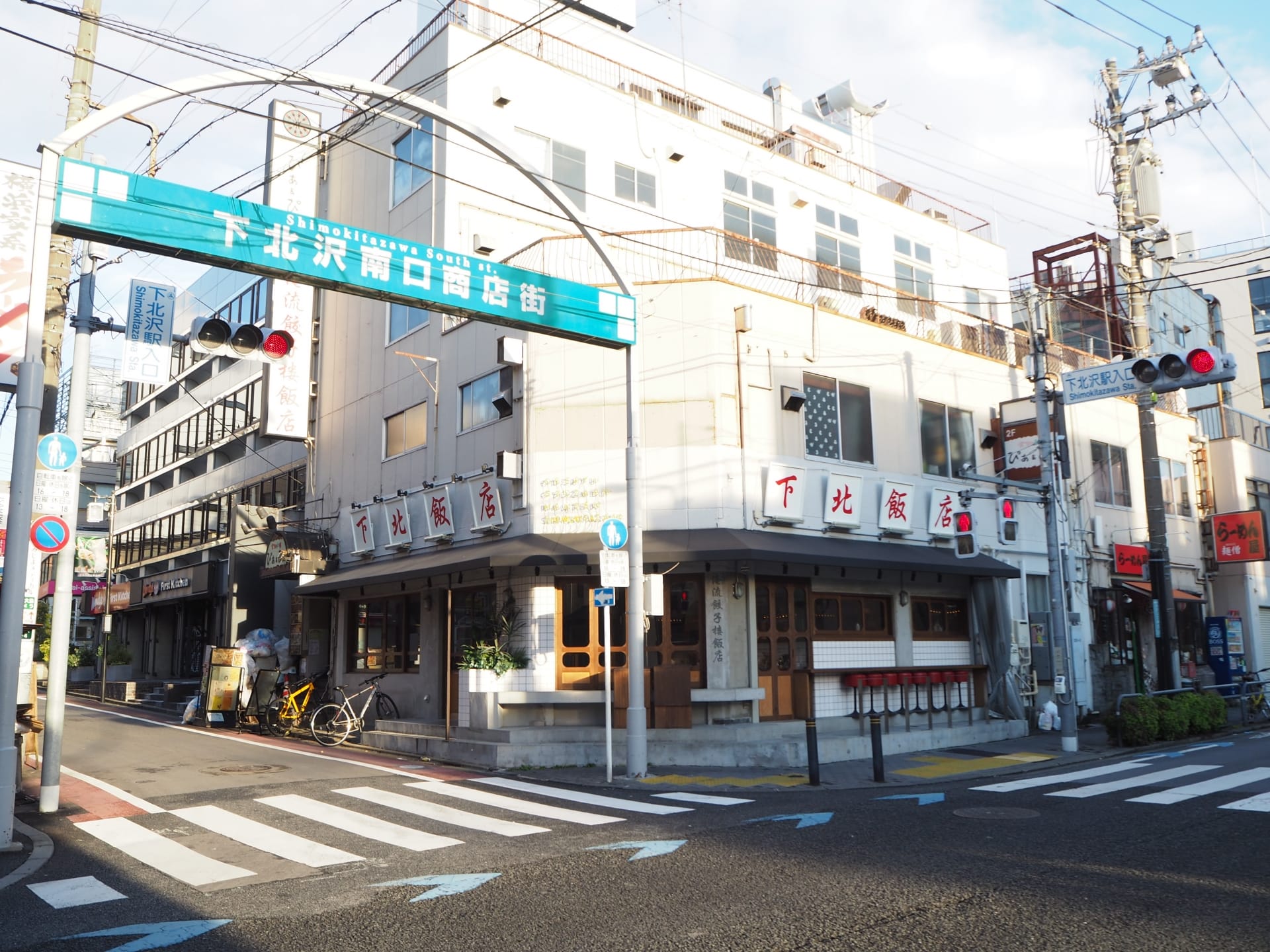 下北沢の街並みには、青い日本語の看板の下にある建物や店舗が特徴です。自転車は左側にあり、交差点は信号機で見渡せます。一方、韓国語教室マンツーマンの広告が、この魅力的な近所のスナップショットに文化的な雰囲気を加えています。.