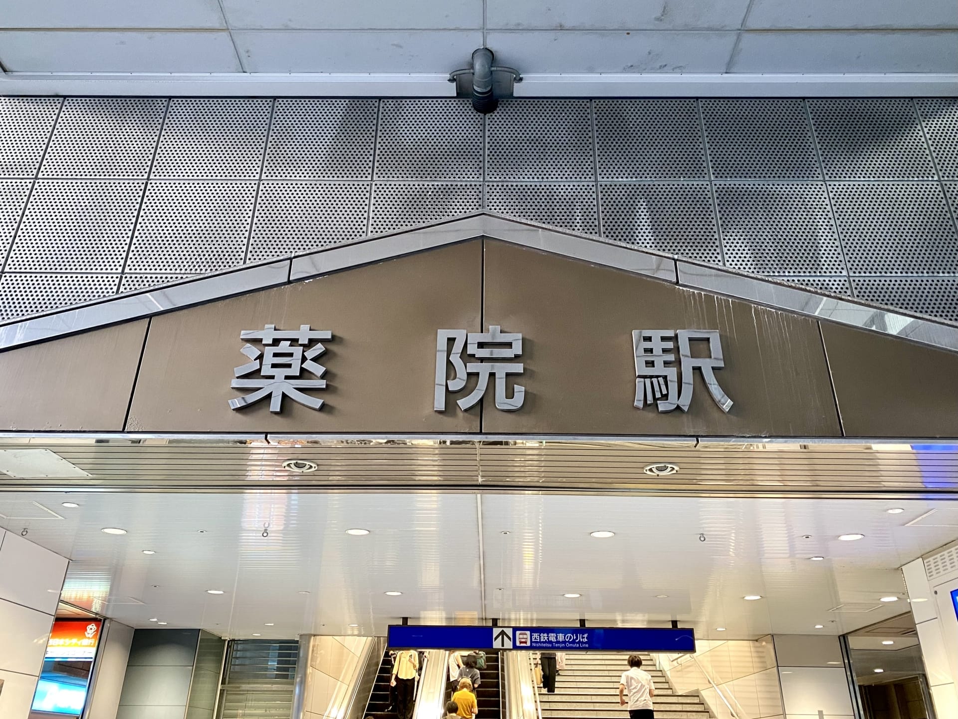 薬院駅の入口の看板の上には日本語の文字が誇らしげに表示されており、その下では人々がエスカレーターで上がっており、おそらく韓国語のマンツーマンレッスンへ向かっている。.