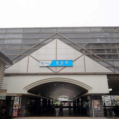 京成駅の外観は、印象的な三角屋根と入口上の青い看板が特徴で、賑やかな都市景観を背景に、経堂に通う人々が帰宅途中に韓国語教室のマンツーマンを探していたような場所を思い出させます。.