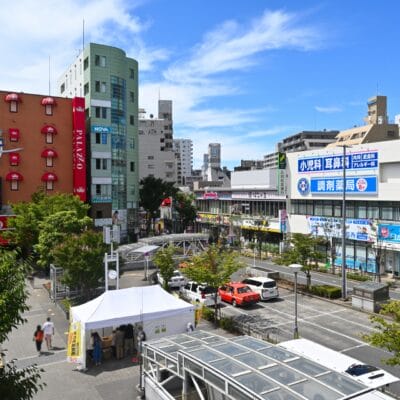 葛西の活気に満ちた都会の街並みの中に、時計と広告のある赤い建物を含む建物がそびえ立っています。前景に緑が美しく映える中、車と白いテントが道路に点在しています。明るく晴れた日には、この活気に満ちた背景が強調され、この地域での韓国語教室マンツーマンのセッションに最適です。.