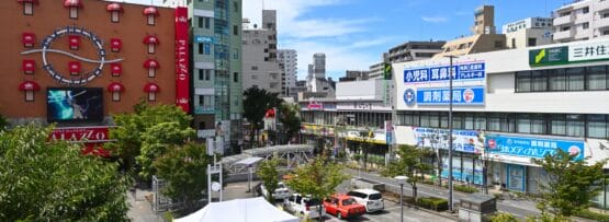 葛西の活気に満ちた都会の街並みの中に、時計と広告のある赤い建物を含む建物がそびえ立っています。前景に緑が美しく映える中、車と白いテントが道路に点在しています。明るく晴れた日には、この活気に満ちた背景が強調され、この地域での韓国語教室マンツーマンのセッションに最適です。.