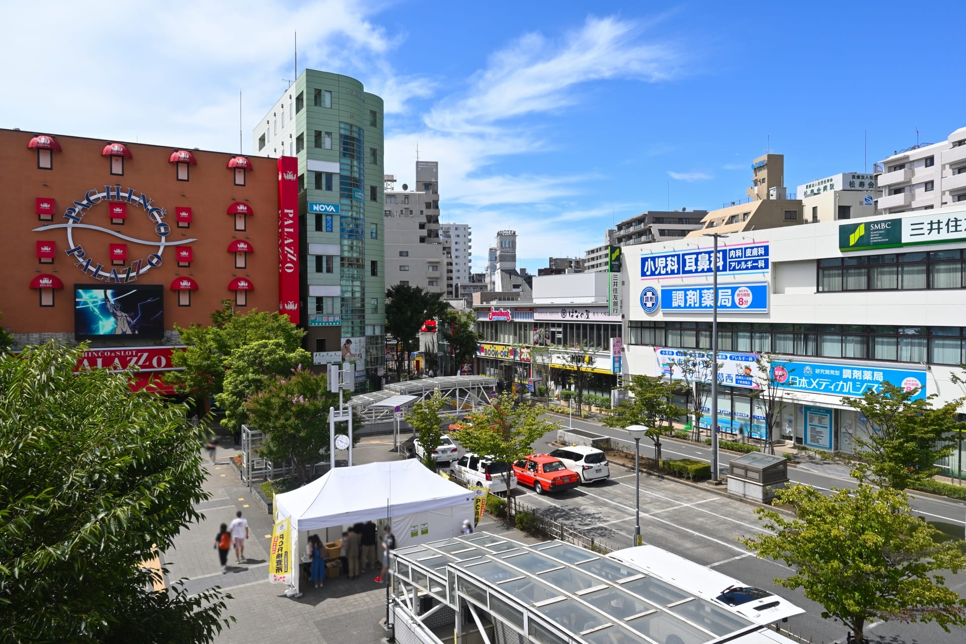 葛西の活気に満ちた都会の街並みの中に、時計と広告のある赤い建物を含む建物がそびえ立っています。前景に緑が美しく映える中、車と白いテントが道路に点在しています。明るく晴れた日には、この活気に満ちた背景が強調され、この地域での韓国語教室マンツーマンのセッションに最適です。.