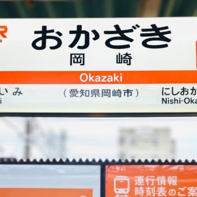 岡崎駅CA52の日本語とローマ字が描かれた駅名標には、愛美と西岡崎への行き先も記載されています。背景がぼやけて駅の屋根がそびえ立っています。近くには韓国語教室マンツーマンもある岡崎を見つけてください。.