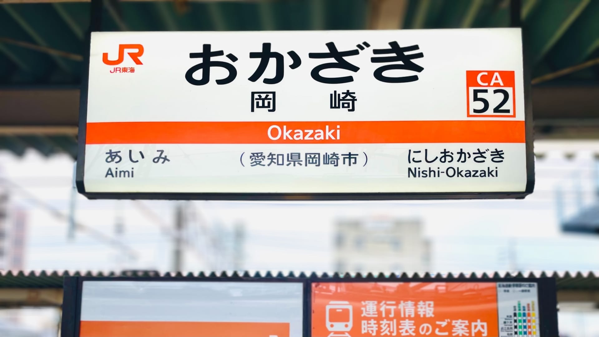 岡崎駅CA52の日本語とローマ字が描かれた駅名標には、愛美と西岡崎への行き先も記載されています。背景がぼやけて駅の屋根がそびえ立っています。近くには韓国語教室マンツーマンもある岡崎を見つけてください。.