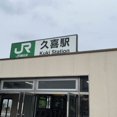 JR久喜駅の入口には、日本語と英語で駅名が誇らしげに掲げられた看板が掲げられています。近くでは、曇り空の下、久喜の繁華街にある韓国語教室マンツーマンのクラスに向かう人が玄関を歩いている。.