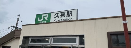 JR久喜駅の入口には、日本語と英語で駅名が誇らしげに掲げられた看板が掲げられています。近くでは、曇り空の下、久喜の繁華街にある韓国語教室マンツーマンのクラスに向かう人が玄関を歩いている。.