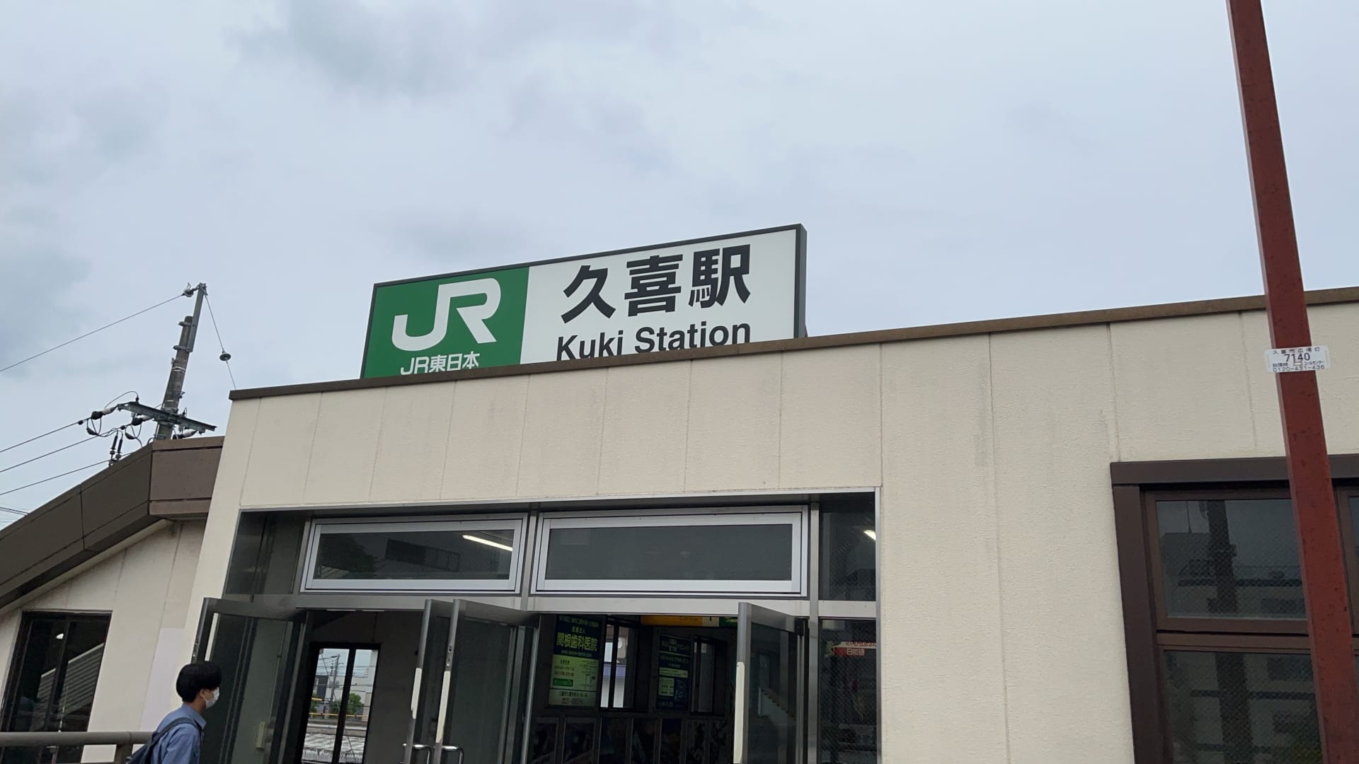 JR久喜駅の入口には、日本語と英語で駅名が誇らしげに掲げられた看板が掲げられています。近くでは、曇り空の下、久喜の繁華街にある韓国語教室マンツーマンのクラスに向かう人が玄関を歩いている。.