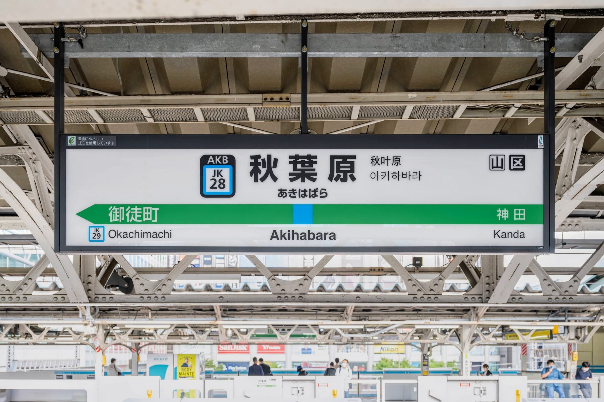 駅のプラットホームは活気に満ち、看板には英語、日本語、韓国語、中国語で「秋葉原」と誇らしげに表示されています。プラットホームの番号は旅行者を案内し、矢印は御徒町と神田を指しています。近くでは、熱心な学習者を待つ韓国語のマンツーマンクラスがあります。.