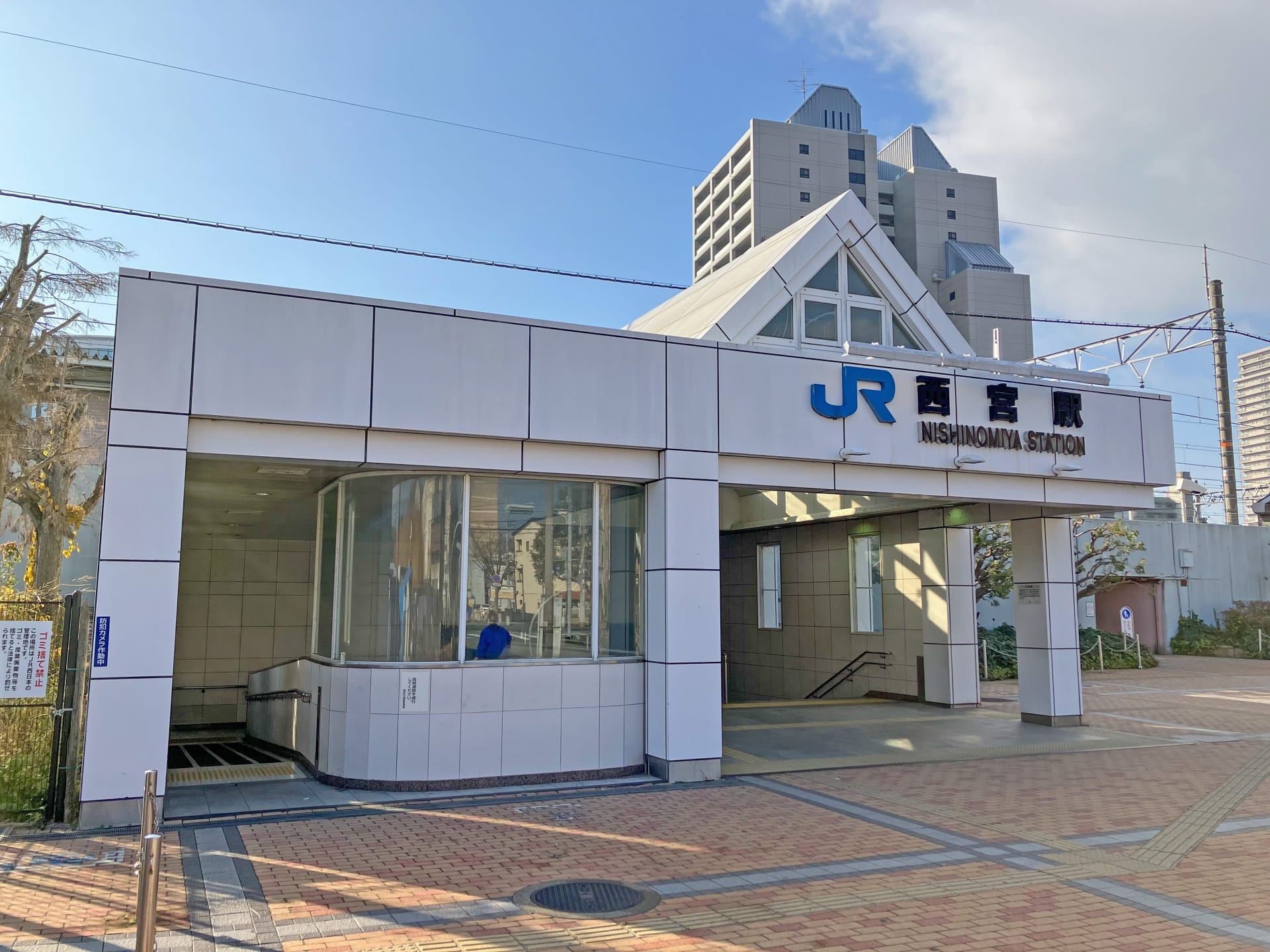 JR西宮駅の入口。一面ガラス張りの白い建物が特徴。曇り空の下、背景に高層ビルが見え、近くにある個人向けの韓国語教室マンツーマンのような活気に満ちた文化と多様な機会を反映しています。.