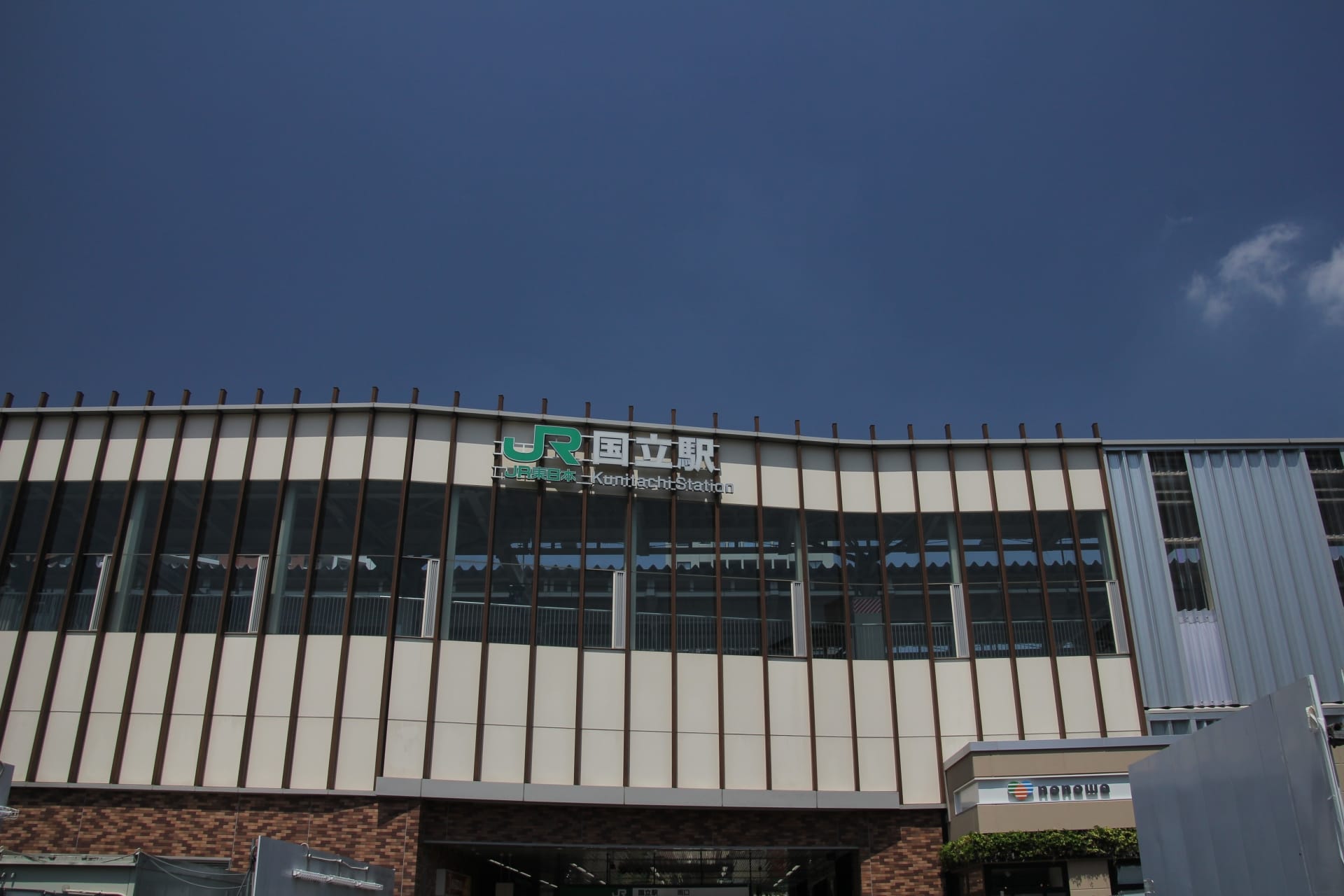 青空を背景に看板とコンビニエンスストアの入り口が特徴的なJR国分寺駅の外観。近くには、個別の韓国語レッスンを提供する国立の人気の韓国語教室マンツーマンがあります。.