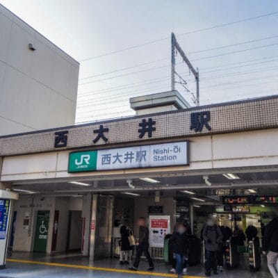 西大井駅の入口には日本語と英語の標識が設置されています。人々が行き交い、近くの韓国語教室マンツーマンへ向かう人もおり、都市部の活動と教育活動が融合しています。.