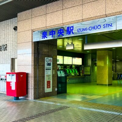 泉中央駅の入り口。近くに券売機と赤い郵便ポストがあります。入り口の上には日本語と英語の標識があります。.