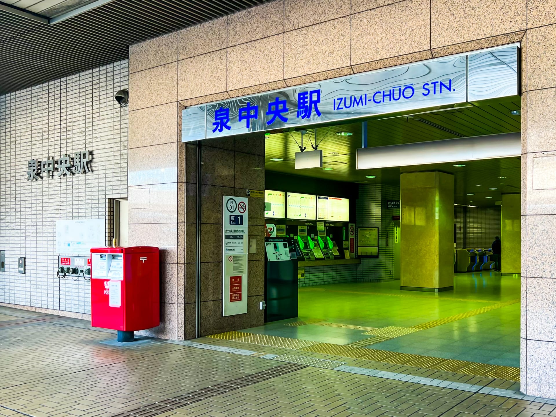 泉中央駅の入り口。近くに券売機と赤い郵便ポストがあります。入り口の上には日本語と英語の標識があります。.