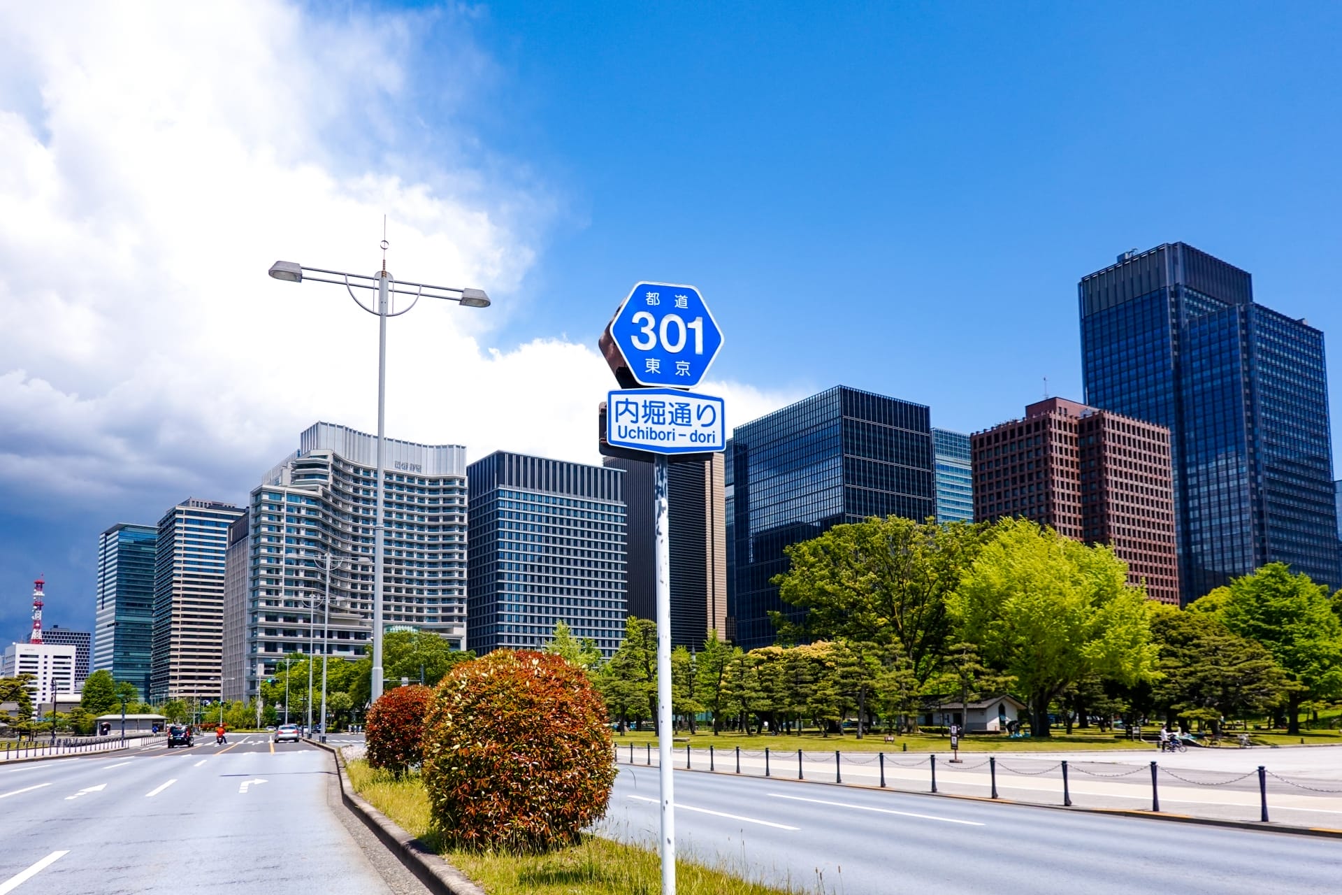 日本の東京にある内堀通り 301 の道路標識は大手町を指しており、部分的に曇った空の下、背景には高層ビルと緑が広がっています。.