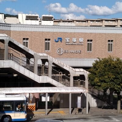 JR高月駅「エキマルシェ」と名付けられた2階建ての建物は、屋外階段で入り口へと続いており、目立つ位置にあります。正面の木々の横にはバスが停まっています。近くには宝塚劇場があり、都会の雰囲気の中で文化的な雰囲気を味わえます。.