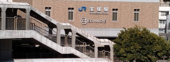 JR高月駅「エキマルシェ」と名付けられた2階建ての建物は、屋外階段で入り口へと続いており、目立つ位置にあります。正面の木々の横にはバスが停まっています。近くには宝塚劇場があり、都会の雰囲気の中で文化的な雰囲気を味わえます。.