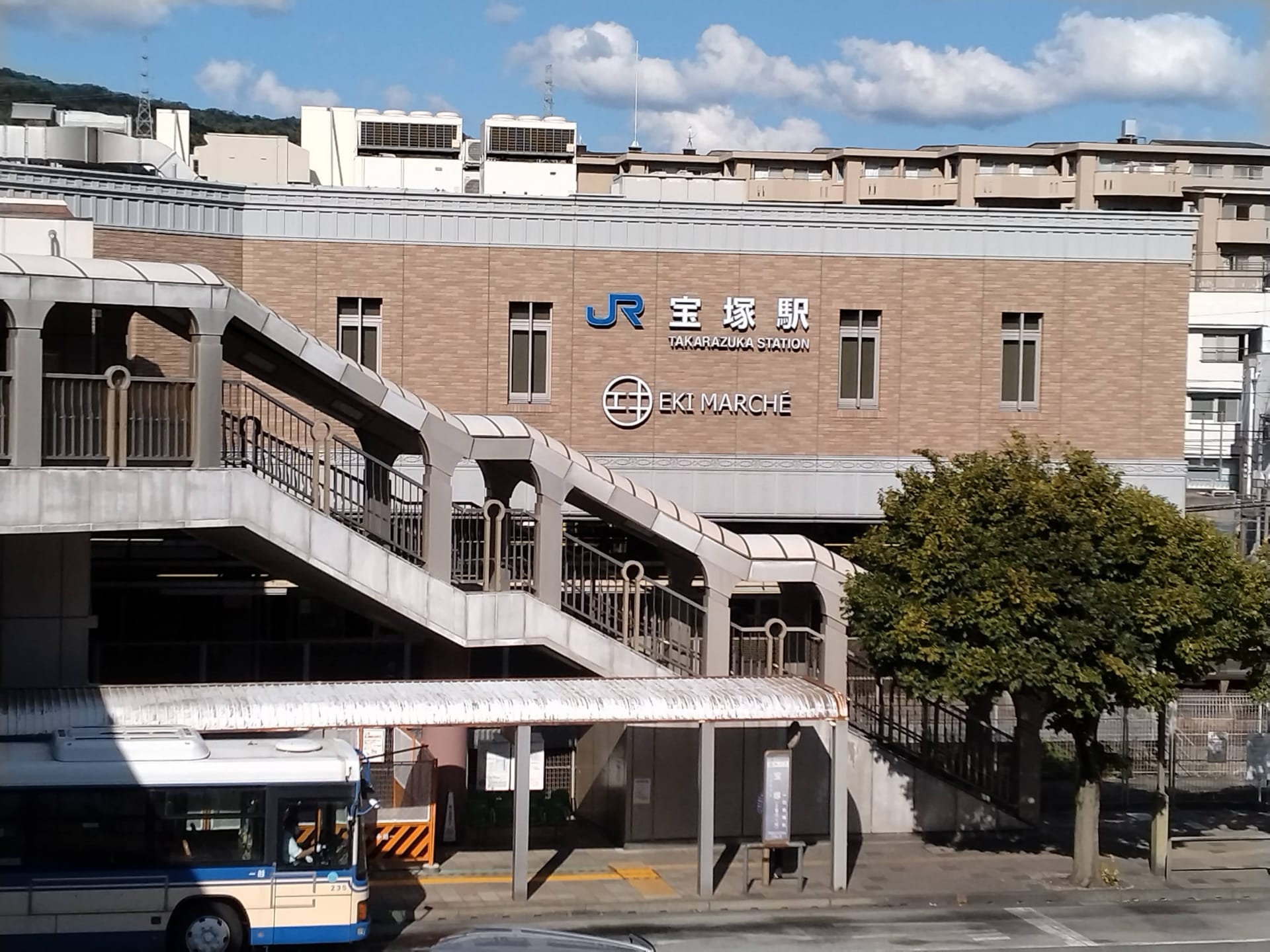 JR高月駅「エキマルシェ」と名付けられた2階建ての建物は、屋外階段で入り口へと続いており、目立つ位置にあります。正面の木々の横にはバスが停まっています。近くには宝塚劇場があり、都会の雰囲気の中で文化的な雰囲気を味わえます。.