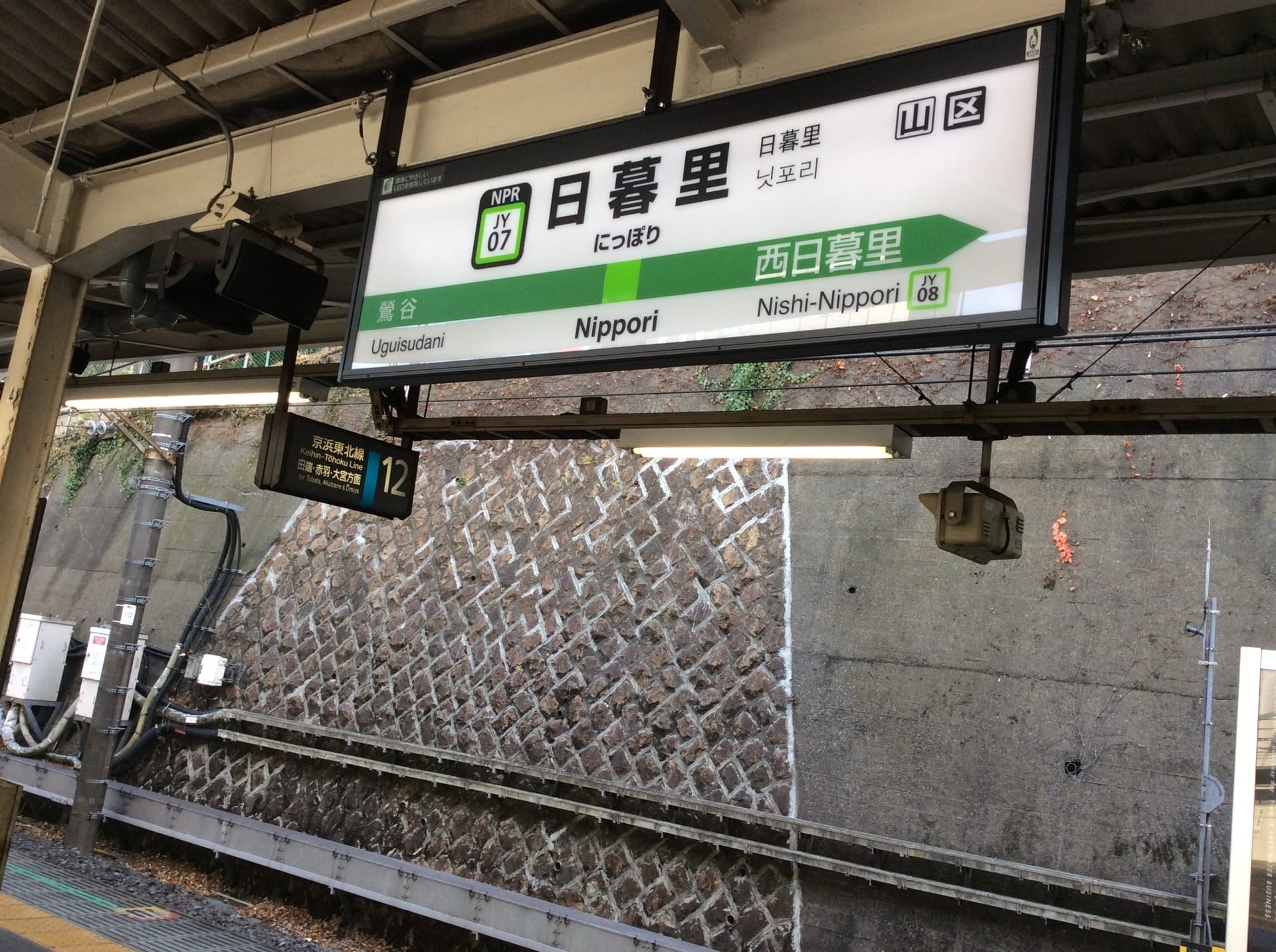 駅のホームには「日暮里」とホーム番号が書かれた看板があり、その背後には石垣が広がっています。柱には防犯カメラが設置されており、賑やかな日暮里の中心地を見守っています。.